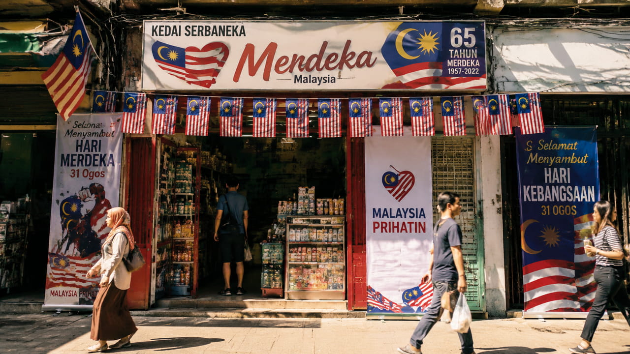 A small retail shop storefront in Kuala Lumpur decorated with red, white, and blue Merdeka-themed posters and bunting flags